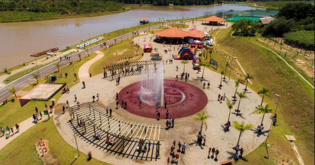 Vista aérea do Parque do Mirim em Indaiatuba com fonte interativa, estruturas de sombra, brinquedos infláveis e lago ao fundo.