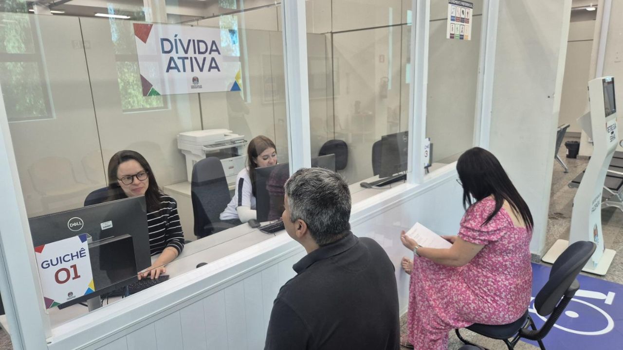 Imagem mostra um guichê de atendimento público com um vidro que separa as duas atendentes das duas pessoas que estão sendo atendidas, um homem vestindo blusa preta e uma mulher com vestido rosa estampado. Todos estão sentados em cadeiras pretas. No vidro tem um adesivo escrito DÍVIDA ATIVA.